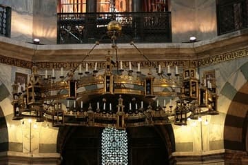 Barbarossa's Chandelier, hanging from the vault in the Palatine Chapel. The huge (4.2-meter diameter) bronze circlet was commissioned by Frederick Barbarossa on the occasion of Charlemagne's canonization. It was created 1165-84 in Aachen and is inscribed with a dedication to Mary from Barbarossa and his wife Beatrix. The candles are lit on special holy days. The chandelier's design represents the Heavenly Jerusalem as envisioned in Revelation, with eight towers instead of the twelve described in Revelation. The dedicatory inscription explains that the deviation from the biblical account was so that the chandelier would fit perfectly into the eight-sided imperial chapel.