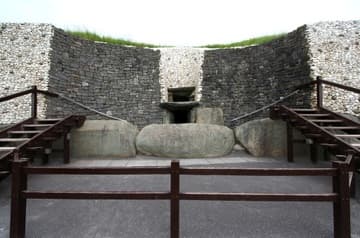 Facade and entrance at Newgrange, a prehistoric passage tomb and ceremonial chamber dating from about 3200 BC. Boyne River Valley, County Meath, Ireland. The facade around the perimeter is made of sparkling white quartz brought from 50 miles away, punctuated by egg-shaped granite stones. The facade has been shaped by archaeologists to allow for visitor access; the dark stones that curve inwards were deliberately chosen to show they are not an original feature of the tomb. Originally, the white facade continued above the round stones, with a narrow recess leading to the entrance. Directly in front of the entrance is the Threshold Stone, which is elaborately carved with spirals, concentric circles and diamond shapes. Their meaning is not known but theories are plentiful. Above the entrance is a "roof box" that allows sunlight to stream in on the winter solstice.