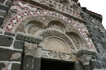 The fascinating facade of the chapel dates from the 12th century. Like Le Puy Cathedral, its multicolor stonework and arches give it an exotic appearance revealing Islamic influences. The stonework of the facade illustrates themes from Genesis and Revelation. The reliefs within the unusual trefoil arch depict the Lamb of God (labeled with AGNUS DEI) in the center, with four Elders of the Apocalypse holding vials of perfume on each side. Above the arch are vines populated with birds and two human figures, which probably represents Creation and the Fall. Note the tiny Green Man masks beneath the feet of each human figure.