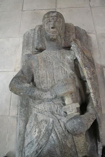 One of ten knightly memorials in the round nave of the Temple Church, London. These were believed to be tombs until the post-WWII restoration revealed no bodies, but only effigy memorials. All the knights are on their back, but are otherwise positioned in different ways: some have their legs extended straight out while others have their legs crossed; some wear tunics over their armor and others wear full-length robes; some clutch their swords, some pray, and some have their arms straight at their sides. One has no effigy at all, but only a trapezoidal sarcophagus lid.