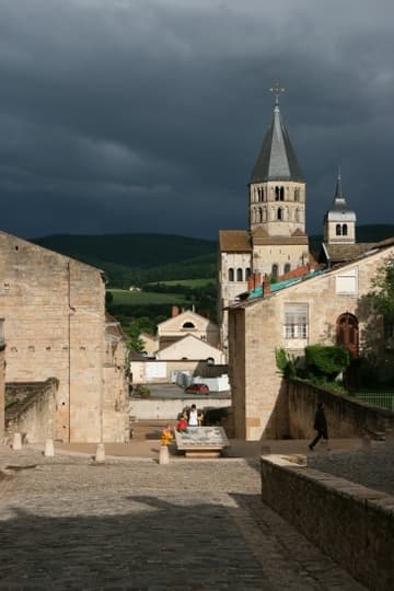 Site of the former Cluny Abbey church (1088-1130), of which only the south transept survives, seen from the west in some dramatic evening light. The semicircular area in the foreground is the beginning of the parvis (an outdoor staircase). This led down to the huge antechurch or porch (1130-90), in which the foundations of pillars can be seen. The sloped-roof stone buildings are the surviving bases of the great west towers, the Barabans. Against a landscape of Burgundian hills in the background is the south transept, topped with the beautiful Clocher de l'Eau-Bénite ("Holy Water Belfry").