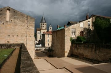 Site of the former Cluny Abbey church (1088-1130), of which only the south transept survives, seen from the west. The area in the parvis (an outdoor staircase). This led down to the huge antechurch or porch (1130-90), in which the foundations of pillars can be seen. The sloped-roof stone buildings are the surviving bases of the great west towers, the Barabans. Against a landscape of Burgundian hills in the background is the south transept, topped with the beautiful Clocher de l'Eau-Bénite ("Holy Water Belfry").